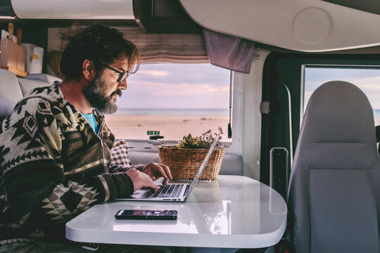 An adult man works on his laptop inside a modern motorhome with a beach and ocean view outside the window, using his phone for a hotspot and data roaming to stay connected and productive