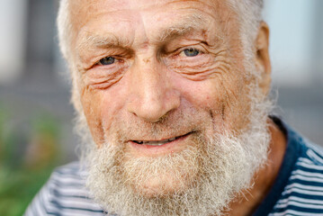 Senior man in his 80s enjoying a relaxed moment outdoors, smiling joyfully with wrinkles, white hair, blue eyes, and a beard, capturing the essence of aging gracefully while focusing on physical 