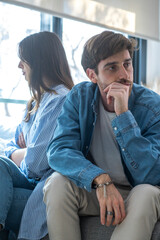 Young millennial couple sitting back to back on a sofa at home, sad and upset, ignoring each other,...