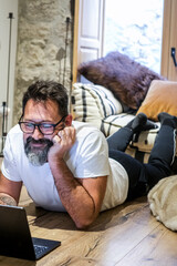 A 50-year-old man is relaxing at home on the floor, lying beside a dog, using a laptop to browse the web and search for information while a fire burns in the background of the living room