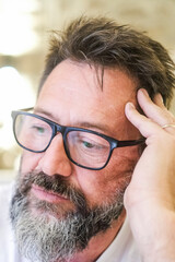 A middle-aged man, about 50 years old, sits alone at home wearing glasses, his face displaying an...