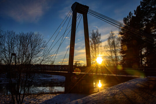 A modern suspension bridge silhouetted against a stunning winter sunset, with sun rays and reflection on a calm river, surrounded by trees and snow.