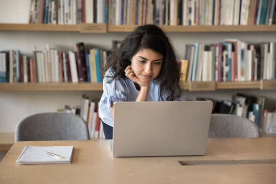 Searching information. Focused young female student engaged in learning process in university library lean at work desk use notebook pc to contact groupmate tutor in chat check data received from book