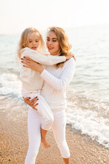 Smiling young mother 30-35 year old holding child girl 5-6 year old over sea at beach wearing knitted sweaters and denim pants outdoor. Motherhood. Summer vacation time.