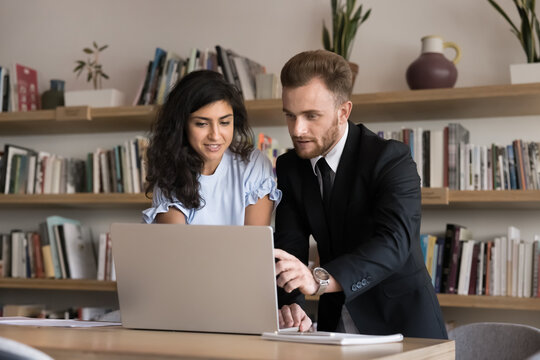 Supportive role of mentor. Interested female student listen to young male teacher assistant showing work details in e-learning app on notebook at library engaged in collaboration with experienced peer