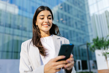 Successful young adult businesswoman using smart phone over business building. Technology lifestyle and business concept