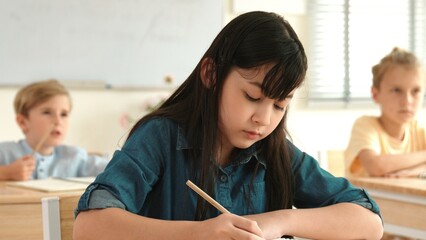 Asian girl writing answer in paper while focus on thinking about theory during test. Diverse smart students taking a note or lecturing while teacher teaching in elementary class. Education. Pedagogy.