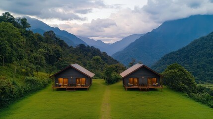 Obraz premium Two cozy cabins nestled in a lush green valley, surrounded by mountains and clouds, offering a serene getaway in nature.