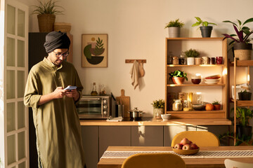 Man wearing traditional clothing standing in kitchen while using cellphone. Modern kitchen adorned with plants, wooden shelves, and various kitchen items