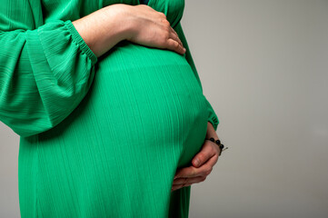 Close-up image of pregnant woman in green dress holding hands near belly, expecting baby. Symbol of...