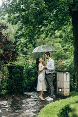 couple standing under a transparent umbrella. A man in a white shirt hugs a woman in a white dress. They are on the street, there are green tree leaves around them.