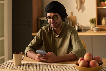 Man wearing turban and glasses sitting at table reading on smartphone with mug of coffee and fruits visible capturing a casual kitchen setting
