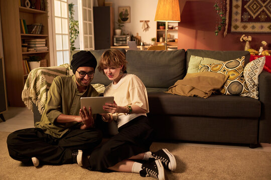Two friends sitting on floor, laughing while viewing something funny on a tablet in cozy living room setting with comfortable furniture and warm lighting, creating a relaxed atmosphere