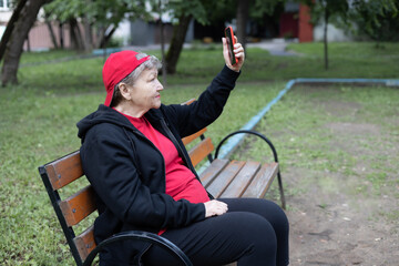 Portrait of happy elderly woman in red cap and sportwear sitting on bench and smiling taking selfies on smartphone on background of green park. Active life of pensioners and adaptation in the modern w