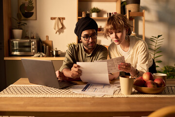 A couple is siting at a kitchen table, reviewing financial documents attentively. Both individuals are focused on paperwork, with a laptop, coffee mugs, and fruit centerpiece visible