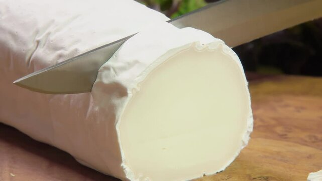 The knife cuts off a piece of Sainte maure de Touraine goat cheese on a wooden board on a background of fresh vegetables. High quality 4k footage