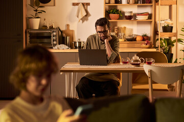 Man in cozy kitchen setting working on laptop with thoughtful expression. Blurred person in foreground using smartphone adding depth and context to scene