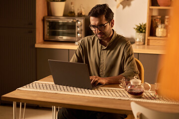 Man in kitchen working on laptop, surrounded by warm lighting and cozy decor, indicating relaxed yet focused atmosphere in home office setting