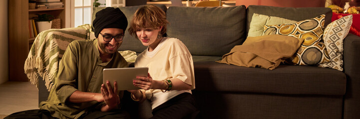 Two friends sitting on floor using tablet in warm and cozy living room environment. Smiling and...