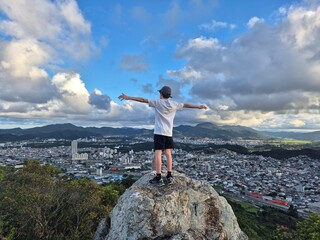Obraz premium menino no alto da montanha com braços abertos observando a vista da cidade 