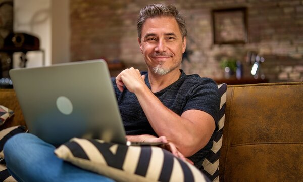 Older man working online with laptop computer at home sitting on couch in living room. Home office, browsing internet. Portrait of happy, mature age, middle age, mid adult man in 50s, smiling.
