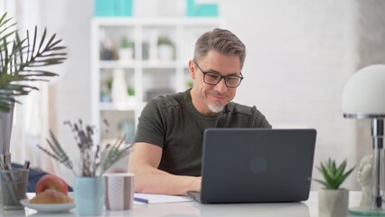 Middle aged casual man typing on laptop, browsing internet, reading email with morning coffee. Mid adult male entrepreneur managing business online. Businessman in glasses working in home office.