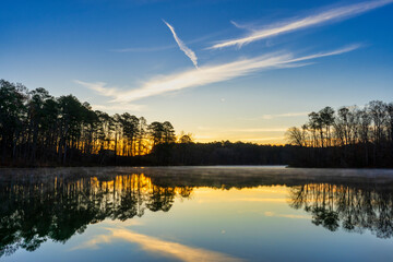 As the sun starts to rise over Chief McIntosh Lake in Georgia a warm glow illuminates the landscape. The silhouetted trees and a brilliant blue sky with wispy clouds are reflected in the calm water.