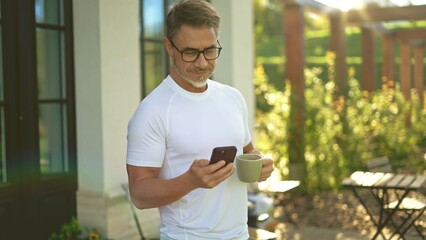 Relaxed mature man standing on terrace in garden drinking coffee checking smartphone in morning sun. Businessman outdoor at home, perfect start of a long business day. Life of successful entrepreneur.