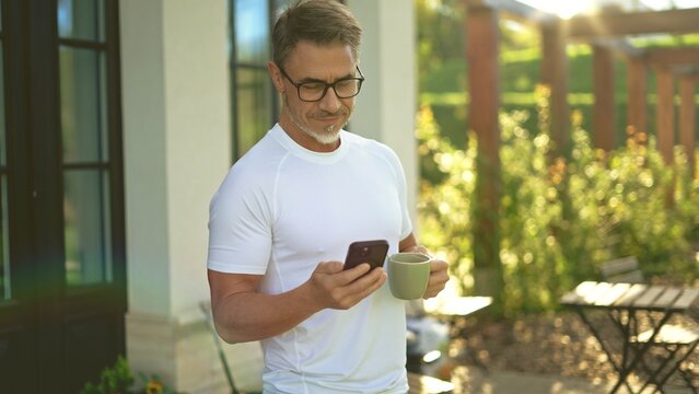 Relaxed mature man standing on terrace in garden drinking coffee checking smartphone in morning sun. Businessman outdoor at home, perfect start of a long business day. Life of successful entrepreneur.