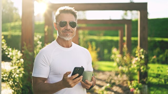 Happy mature man using smartphone drinking coffee in garden during beautiful sunrise. Portrait of senior male in sunglasses enjoying morning moment of relaxation on home terrace. , smiling .