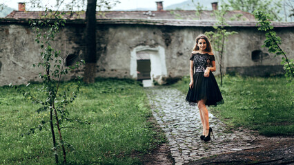The girl smiles and looks at the camera. Around it there is green grass and several young trees. The building in the background looks old and abandoned,
