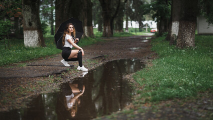 a girl squatting near a puddle on a forest path. She is holding a black umbrella in her hands. The...
