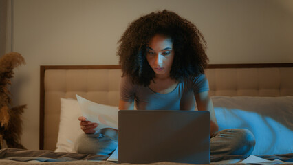 African American woman businesswoman working from home paperwork laptop computer overtime night...