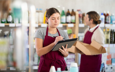 Female manager makes notes on an electronic tablet - conducts an inventory. In background, a male salesperson places goods on shelves