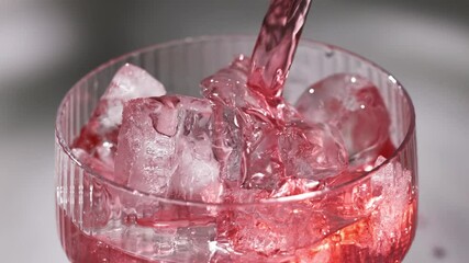 fresh red drink pouring into a cocktail glass with ice cubes on a gray sunny restaurant tabletop, slow motion, zoom in