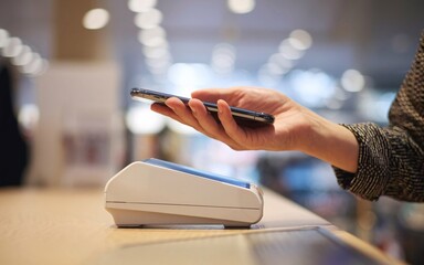 Close up shot of young woman paying contactless on a POS in a shop. She is using her phone.