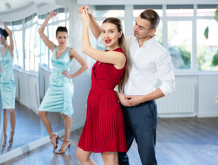 Expressive young adult woman in elegant vibrant red dress dancing passionate bachata with male partner during dance class for adults led by female instructor..