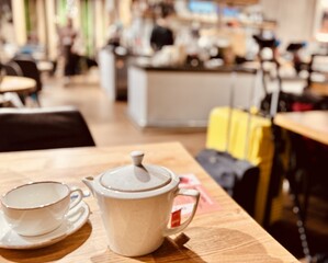 Teapot with green tea and mug with saucer in white color are on table in the restaurant. In the background is yellow suitcase with black metal crutches of vacationing passenger