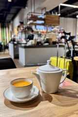 Teapot with green tea and mug with saucer in white color are on table in the restaurant. In the background is yellow suitcase with black metal crutches of vacationing passenger