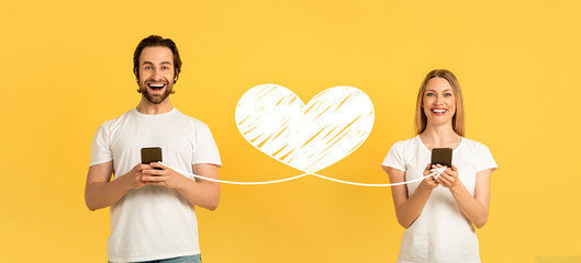 A couple stands together, smiling joyfully while holding their smartphones. They are celebrating St. Valentine's Day by sending affectionate messages to each other.
