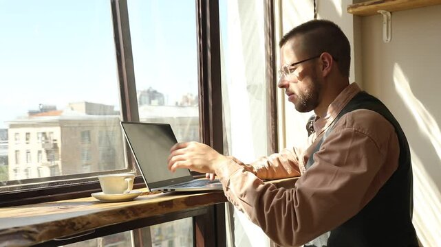It man with glasses by the window working at a computer on a sunny day