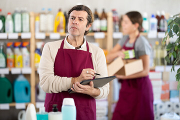Experienced household goods store owner using tablet to manage inventory of cleaning supplies and...