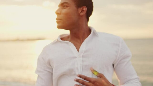 A young man wearing a partially unbuttoned white shirt dances on a beach as the sun sets behind him.