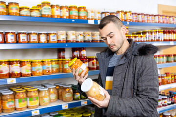 Male shopper selects glass jar of sauerkraut at a grocery store. Man shopper reading expiration date on product label