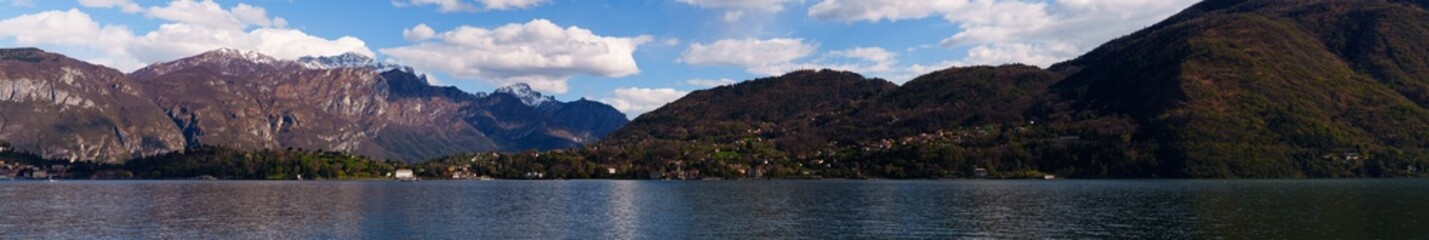 Obraz premium Wide panoramic scene of mountains and Como lake with hillside village nestled at the foot of the hills. Clear blue sky with scattered clouds add to peaceful and scenic atmosphere, Italy