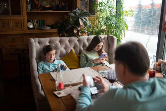 Family sitting at a table in a cafe, everyone looking at their smartphone. Children with their father waiting for their order in a restaurant, brother, sister and father surfing the Internet via mobil