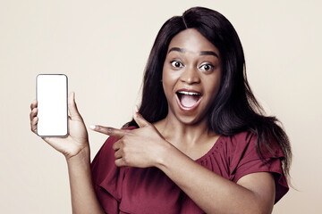 Naklejka premium Excited african american young woman in casual outfit pointing at cellphone with white empty screen, recommending newest mobile application, mockup, beige studio background, copy space