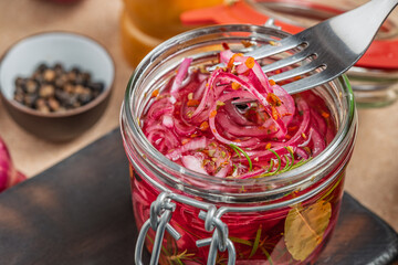 Pickled red onions in a glass jar with spices and herbs. A fork lifts a portion, showcasing the vibrant color and texture. Close-up, rustic kitchen setting.