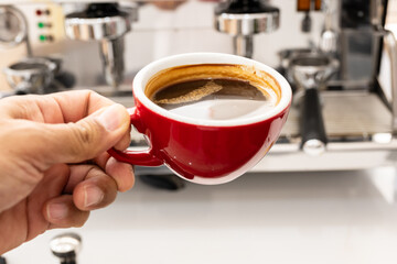Barista hand holding red cup of americano coffee on coffee machine background for breakfast.Top view hot black coffee in restaurant make by barista on best of menu in the coffee shop.Refresh menu.