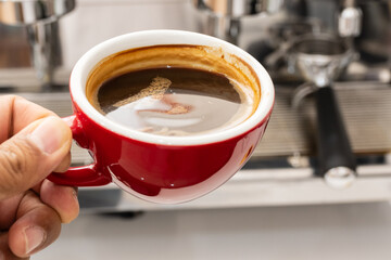 Barista hand holding red cup of americano coffee on coffee machine background for breakfast.Top view hot black coffee in restaurant make by barista on best of menu in the coffee shop.Refresh menu.